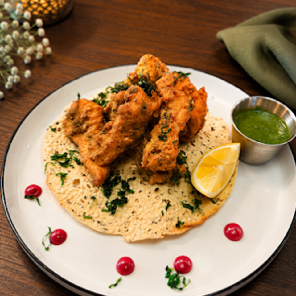 Crispy golden chicken over creamy hummus, garnished with herbs, lemon wedge, green sauce, and pomegranate seeds.