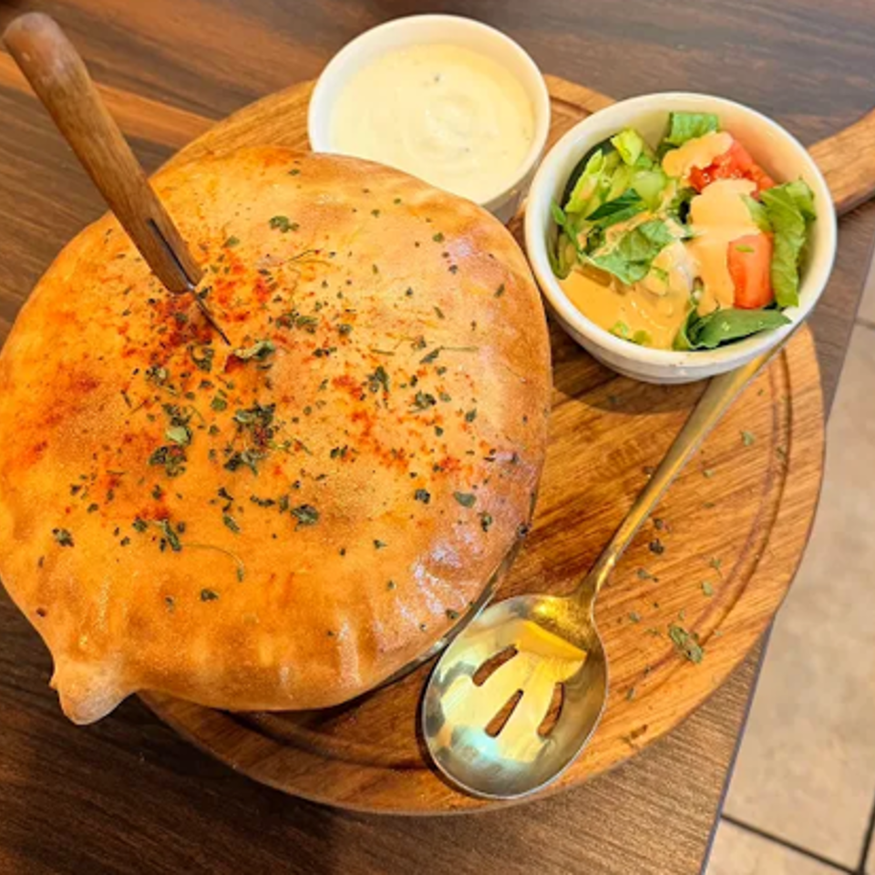 Round herb-seasoned flatbread served on a wooden board with creamy dipping sauce, salad, and a spoon.