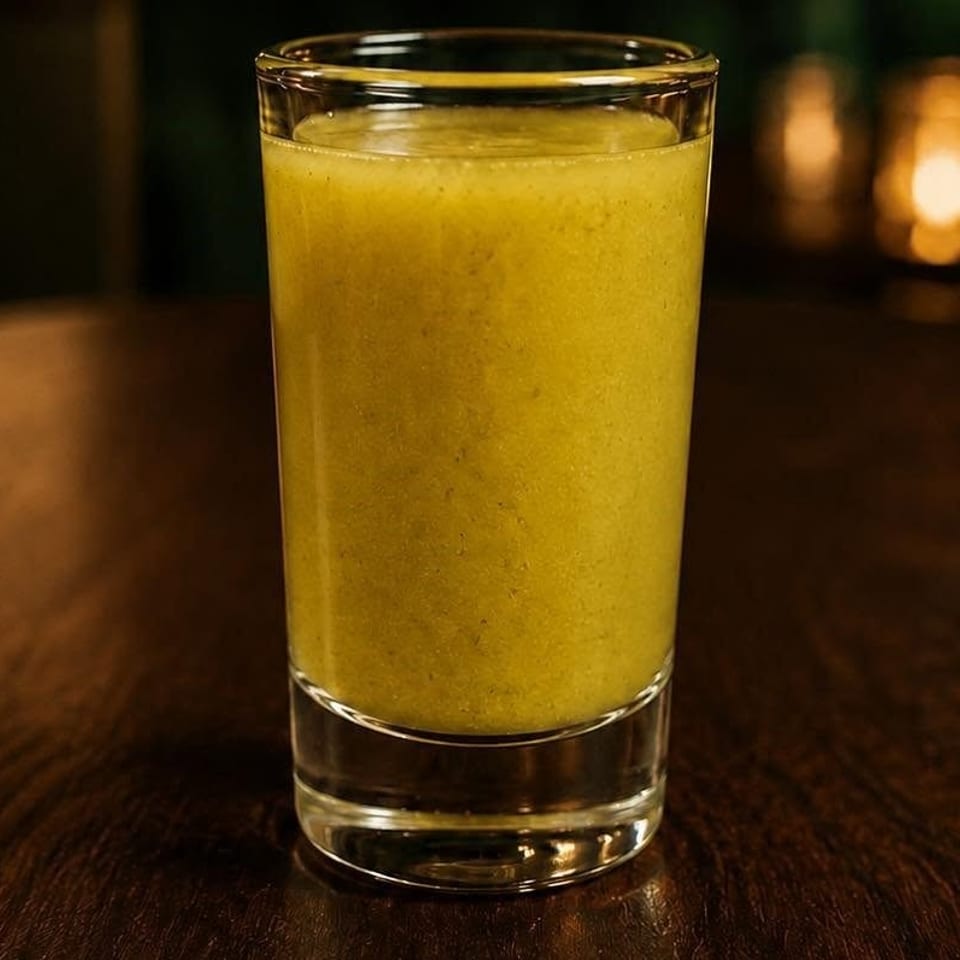 Tall glass of yellow blended drink on a dark wooden table, with warm candlelight glowing in the background.