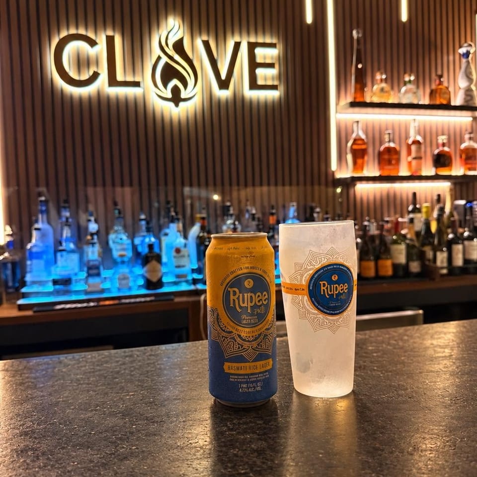Rupee lager can and branded glass on a bar counter, with illuminated liquor shelves and the Clove sign behind them.